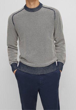 Muž nosící světle šedý svetr s texturou pletení s námořnicky modrým lemováním a tmavě modré kalhoty, ruce v kapsách, na jednobarevném pozadí.