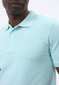Polo in un azzurro chiaro, realizzato in tessuto texturizzato, con una pattina a tre bottoni e un logo ricamato di un uccello sul petto.