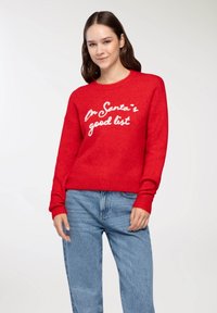 Czerwony sweter z prążkowanymi mankietami i dołem, z białym haftowanym napisem "Na liście dobrych uczniów Świętego Mikołaja". Noszony z jasnoniebieskimi dżinsami.