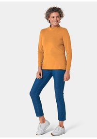 Langärmliges, senfgelbes Oberteil mit einem halben Rollkragen, kombiniert mit blauen Jeans und weißen Sneakers. Weicher Stoff, schlanker Schnitt, minimale Details.
