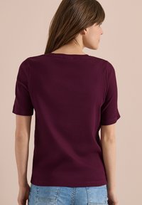 Femme aux longs cheveux bruns portant un t-shirt côtelé à manches courtes, de couleur violet foncé, et un jean en denim bleu clair, dos à la caméra.