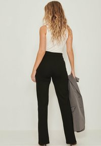 Mujer con cabello rubio ondulado, vistiendo una blusa sin mangas blanca y pantalones negros de cintura alta, sosteniendo una chaqueta gris, de pie contra un fondo sencillo.