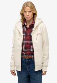 Femme blonde portant un sweat à capuche crème zippé au-dessus d'une chemise en tartan rouge, un jean bleu et une ceinture tressée marron, se tenant contre un fond blanc.