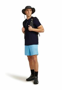 Blue athletic shorts, dark shirt, black hiking shoes, and a dark wide-brimmed hat. The person carries a black backpack with adjustable straps.