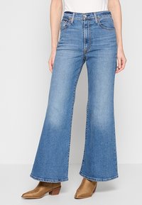 Personne portant un jean bleu taille haute à jambes larges et des bottines marron clair, debout devant un fond clair uni.