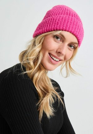 Gorro de lana tejido en rosa con una textura acanalada, se asienta ajustadamente en la cabeza. La persona que lo lleva tiene cabello rubio largo y ondulado y está vestida con una blusa negra.