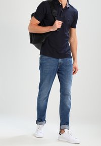 Námořnické polo tričko, modré džíny a bílé tenisky. Model nese černou tašku. Klasický střih, ležérní design, středně těžká látka.