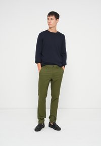 Camisa de manga longa azul-marinho, calças chinos verde-oliva e ténis pretos. Modelo está de pé contra um fundo branco com as mãos nos bolsos.