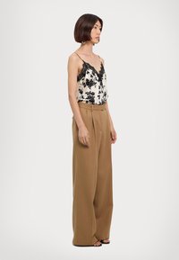 Black lace-trimmed, floral-patterned camisole paired with high-waisted, loose-fitting, tan trousers. Model stands side view on a neutral background.