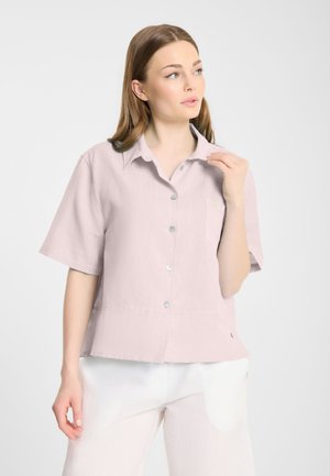 Kobieta o jasnobrązowych włosach, ubrana w luźną, pastelowo-różową koszulę na guziki z krótkim rękawem oraz białe spodnie, patrząca w bok.