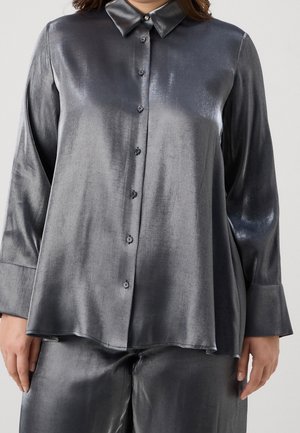 Kobieta ubrana w luźną, błyszczącą, ciemnoszarą koszulę na długi rękaw z guzikami i pasujące spodnie, stojąca na jednolitym tle.