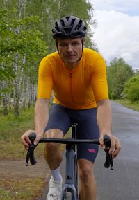 Un cycliste portant un maillot orange vif à manches courtes et un short noir ajusté, pédalant sur un vélo gris sur une route bordée d'arbres.