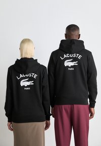 Black hoodies featuring a white crocodile logo and "LACOSTE PARIS" text on the back. The fabric appears soft with a relaxed fit.