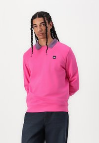 Sudadera rosa con un cuello gris, mangas largas, escote redondo y un pequeño logo negro en el pecho. Color sólido con una textura suave.