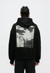 Felpa nera con cappuccio e una grande stampa grafica monocromatica sulla schiena, che presenta un design montano e il logo "Reebok" nella parte superiore.