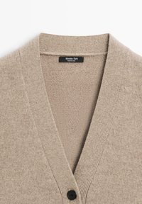 Beżowy dziergany kardigan z dekoltem w serek, wykonany z fakturowanej tkaniny, z jednym czarnym guzikiem z przodu. Etykieta widoczna wewnątrz.