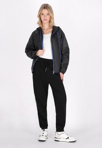 Giacca leggera nera con dettagli blu, abbinata a pantaloni jogger neri e scarpe da ginnastica bianche. La giacca presenta un cappuccio e chiusura con cerniera.