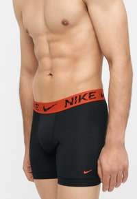 Buste masculin portant un boxer noir Nike avec une ceinture rouge et des logos Nike, debout sur un fond blanc uni.