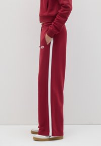 Pantalones de chándal rojos con rayas laterales blancas, hechos de una tela suave, con una cinturilla elástica y bolsillos, combinados con zapatillas blancas.