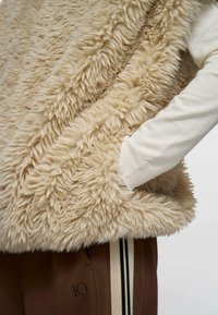 Chaleco de piel sintética beige con una textura suave y esponjosa. Cuenta con un cuello alto y bolsillos laterales. Se lleva sobre una camiseta de manga larga color crema y pantalones marrones.