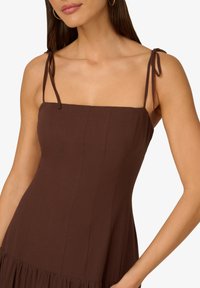 Hnědé šaty s tenkými zavazovacími ramínky, čtvercovým výstřihem, přiléhavým živůtkem, svislými švy a rozšířeným volánkovým lemováním. Hladká textura látky.