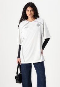 Hvit kortermet skjorte med rutete mønster, grå accenter, og en rund logo. Kombinert med mørke, vide jeans og en svart håndveske.