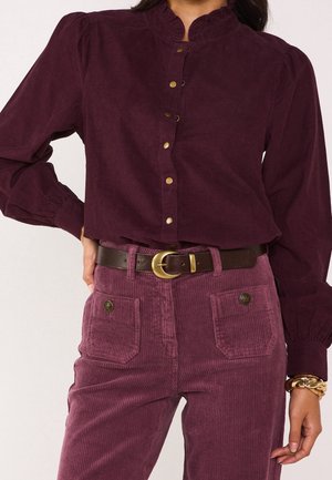 Femme portant un chemisier prune à boutons avec des manches bouffantes et un pantalon en velours côtelé assorti avec des poches avant, une ceinture marron et des bracelets dorés.