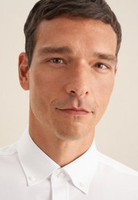 Camicia bianca con colletto classico, realizzata in un tessuto testurizzato e con piccoli dettagli a bottoni sul colletto. Design minimalista.