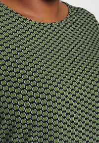 Närbild av nacke och övre bröst som bär en svart skjorta med ett upprepande mönster av gröna ovala former med vit kontur.