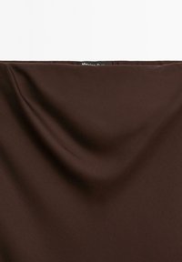 Tessuto marrone scuro con pieghe morbide e una piccola etichetta nera parzialmente visibile al bordo superiore.