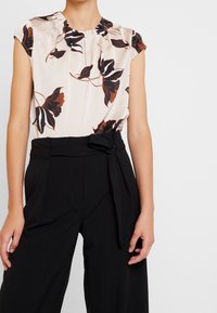 Ärmlös beige blus med svart och brun blommönster instoppad i högmidjade svarta byxor med knytbälte och veck.