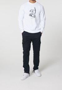 Uomo che indossa una felpa bianca con grafiche di coniglio dei cartoni animati, pantaloni cargo neri e sneaker bianche, in piedi con le mani nelle tasche su uno sfondo bianco.