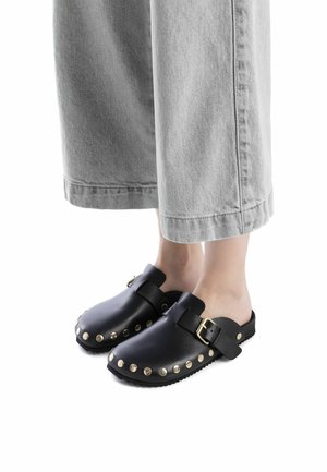 Zuecos de cuero negro con forma redondeada, acentos de tachuelas doradas alrededor de la zona de los dedos y una correa con una hebilla metálica.