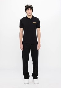 Črna polo majica z zlatim logotipom, v kombinaciji s črnimi hlačami z belimi šivi in belimi supergami. Model nosi z vzorci oblečeno kape.