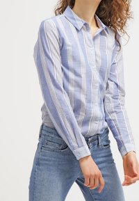 Hellblaue Button-Up-Bluse mit weißen vertikalen Streifen, Kragen, langen Ärmeln, aus Baumwolle oder Leinen, kombiniert mit Jeans in mittlerer Waschung.