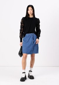 Schwarzer Pullover mit durchsichtigen, floral bestickten Ärmeln, kombiniert mit einem blauen Denim A-Linien-Rock. Das Model hält eine schwarze Tasche und trägt klobige schwarze Schuhe.