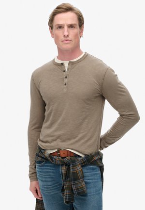 Camisa henley de mangas largas color marrón claro con tres botones negros, combinada con jeans azul y una camisa de cuadros atada a la cintura.