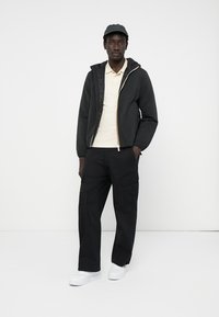 Sobrecamisa negra tipo bomber con forro acolchado, cremallera al frente, camiseta polo beige clara, pantalones cargo negros y zapatillas blancas, complementado con una gorra.