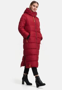 Rode geïsoleerde lange jas met een capuchon, horizontale quiltstructuur, rits aan de voorkant, zijzakken en accenten in oranje-bruin. Gedragen met zwarte enkellaarsjes.