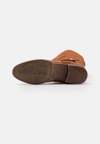 Botte chelsea en daim marron avec une semelle en caoutchouc texturée, un bout arrondi et une petite bride décorative sur le côté. Talon plat.