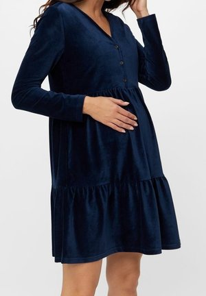 Kobieta w ciąży w ciemnoniebieskiej, długorękawowej sukience ciążowej na guziki, z ręką spoczywającą na brzuchu, stojąca na jednolitym tle.