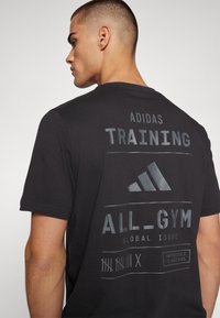 Camiseta de algodón negra con el logo de Adidas, "TRAINING" y "ALL_GYM GLOBAL ISSUE" en impresión gris reflectante en la parte posterior.