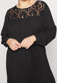 Svart klänning med spetstopp i blommönster, lång ärm och midjeband för justering, tillverkad av mjukt tyg.