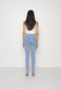 Donna con lunghi capelli scuri, che indossa jeans azzurri, una maglia bianca senza maniche e sandali blu con tacco, in piedi con la schiena rivolta verso di noi.