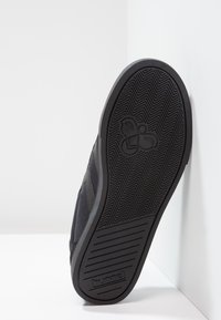 Sapatilha preta com sola de borracha texturizada, apresentando um padrão em zigzag e um logótipo. Parte superior feita de tecido suave com linhas de design elegantes.