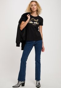 Czarna koszulka z złotym napisem "C'est la vie", w połączeniu z niebieskimi spodniami w dzwony i metalicznymi botkami na obcasie, trzymająca czarną kurtkę.