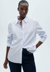 Striped button-up shirt in light blue, made of cotton. Features a collar, long sleeves, and a relaxed fit. Worn with dark jeans.