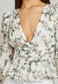 Blusa floreale con scollatura a V profonda, maniche lunghe a sbuffo, vita arricciata e orlo a peplo. Presenta foglie verdi e fiori rosa su uno sfondo crema.