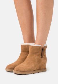 Bottines en daim marron avec une doublure en fourrure douce et une fermeture éclair latérale, dotées d'un talon compensé et d'une semelle en caoutchouc pour une meilleure adhérence.