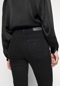 Černé džíny z denimu s čtvercovými ozdobnými prvky, klasickým designem s pěti kapsami a koženou visačkou na pase. Tkanina má hladkou strukturu.